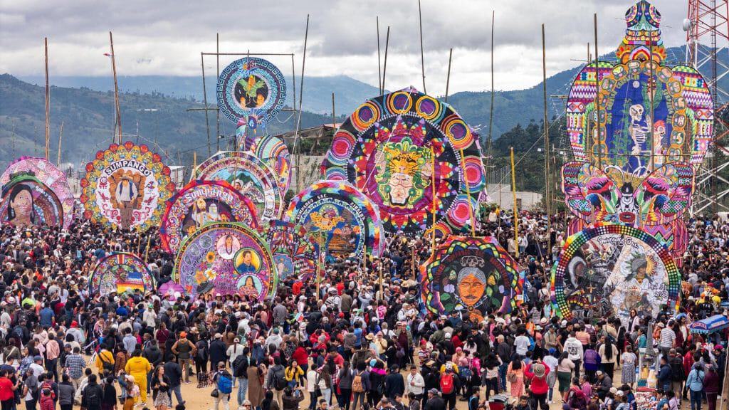 Coloresidas estructuras de papel paja en un festival tradicional con multitud de visitantes en un paisaje montañoso, resaltando la cultura de las festividades populares mexicanas y las expresiones artísticas indígenas. – SoyMigrante.com REVISTA