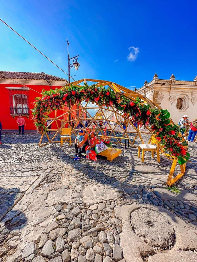 Colorido altar de Día de Muertos en una plaza histórica, adornado con flores y estructura de madera, celebrando la cultura mexicana en un ambiente festivo y vibrante.