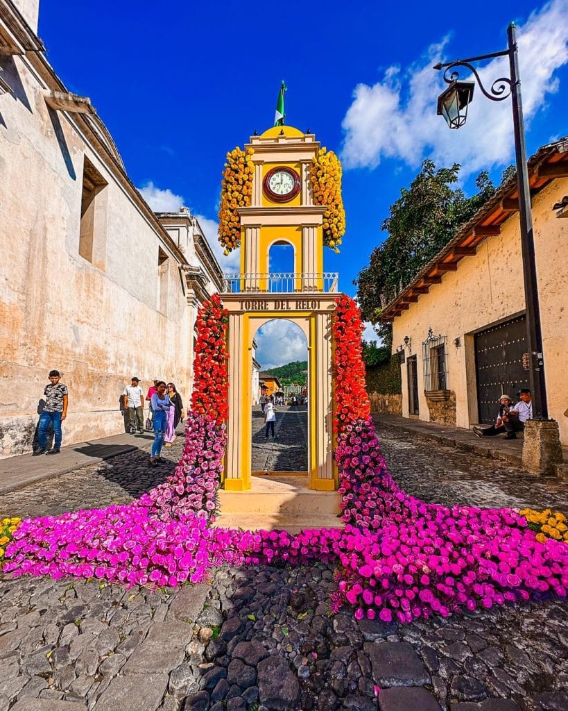 Alta calidad de la Torre del Reloj de Antigua Guatemala decorada con flores vibrantes, representando patrimonio y turismo en Guatemala, ideal para revista de migrantes y cultura.