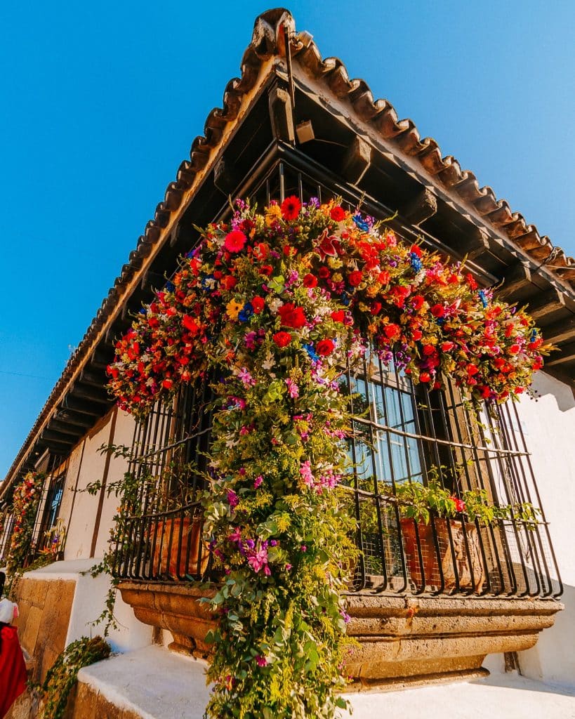 Colorida fachada de casa colonial decorada con flores en el balcón, en un día soleado, reflejando la cultura y belleza de la arquitectura tradicional mexicana.