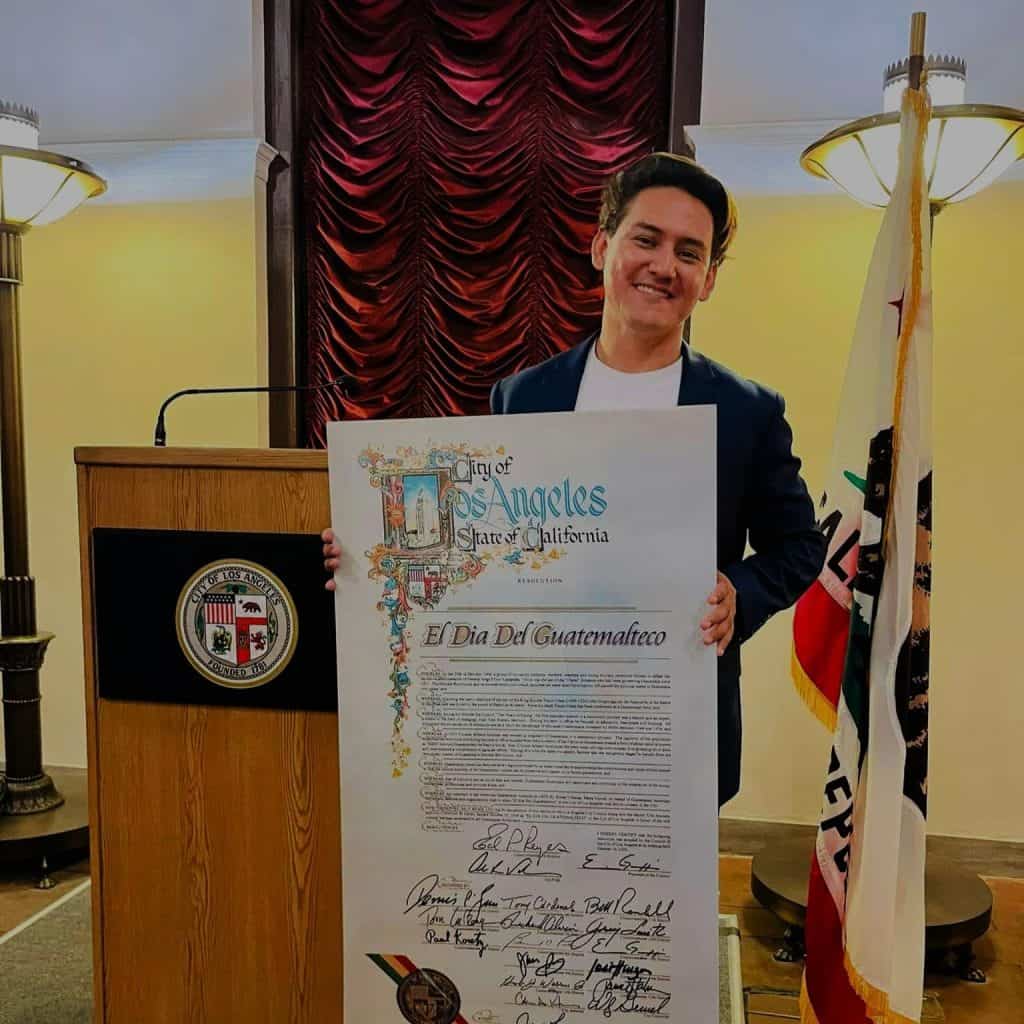 Guatemalteco con resuelto en la ciudad de Los Ángeles, sosteniendo un cartel con la declaración oficial del "Día del Guatemalteco". – SoyMigrante.com REVISTA