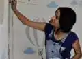 Pintora muralista adolescente creando arte en la pared con aerógrafo en un mural colorido con nubes y globo aerostático, promoviendo cultura y arte en la comunidad. – SoyMigrante.com REVISTA