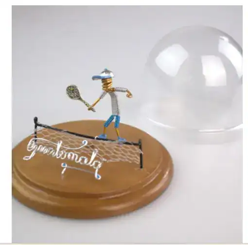 Un pequeño muñeco de alambre con atuendo de verano y raqueta de tenis, decorado en una base de madera, junto a una esfera de cristal, representando creatividad y trabajo artesanal. – SoyMigrante.com REVISTA