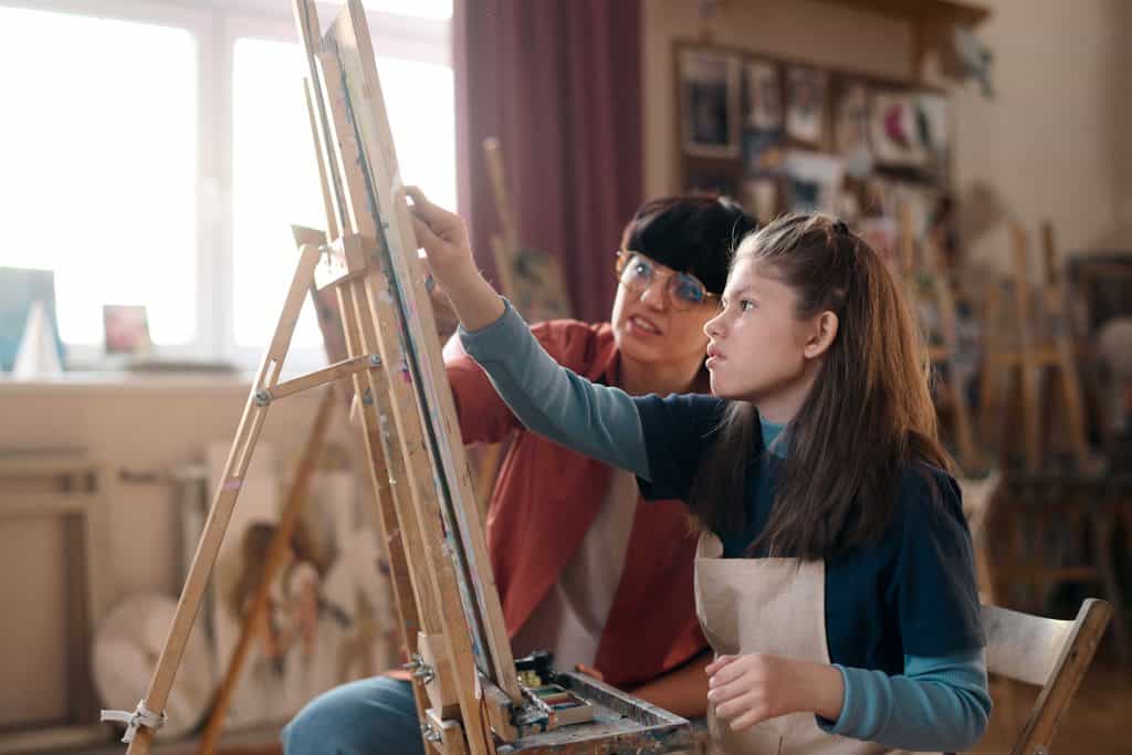 Joven artista y su instructora pintando en un taller de arte para migrantes en un espacio de creatividad y aprendizaje. – SoyMigrante.com REVISTA
