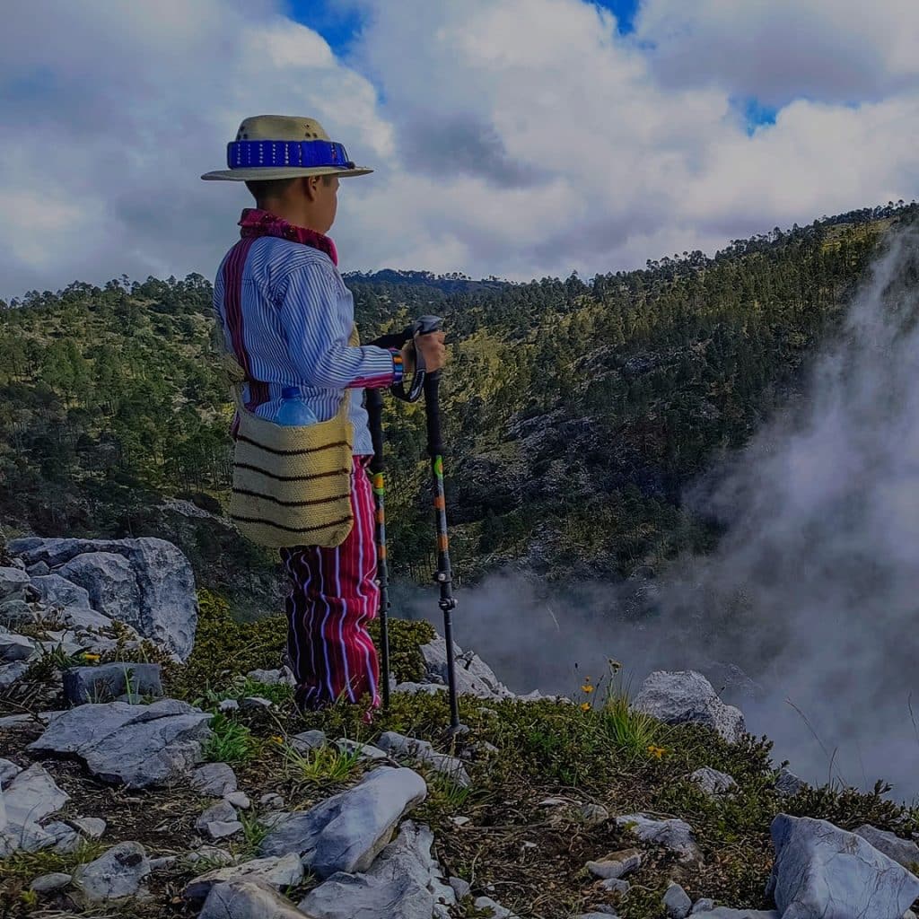 Niña indígena observando un géiser en un parque natural, vestida con ropa tradicional colorida y sombrero de ala ancha, admirando la belleza natural y la cultura ancestral en un entorno montañoso.