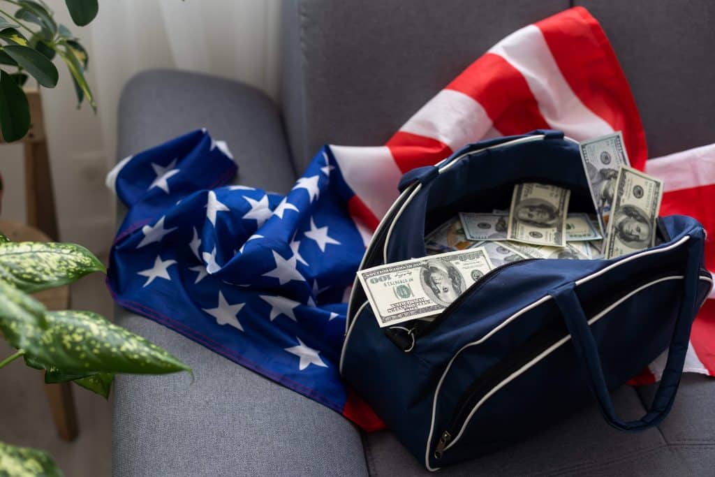Billetes de dólar en maleta con bandera de Estados Unidos y bandera estadounidense en sofá, simbolizando migración, dinero y oportunidades en América, tema destacado en SoyMigrante.com REVISTA. – SoyMigrante.com REVISTA