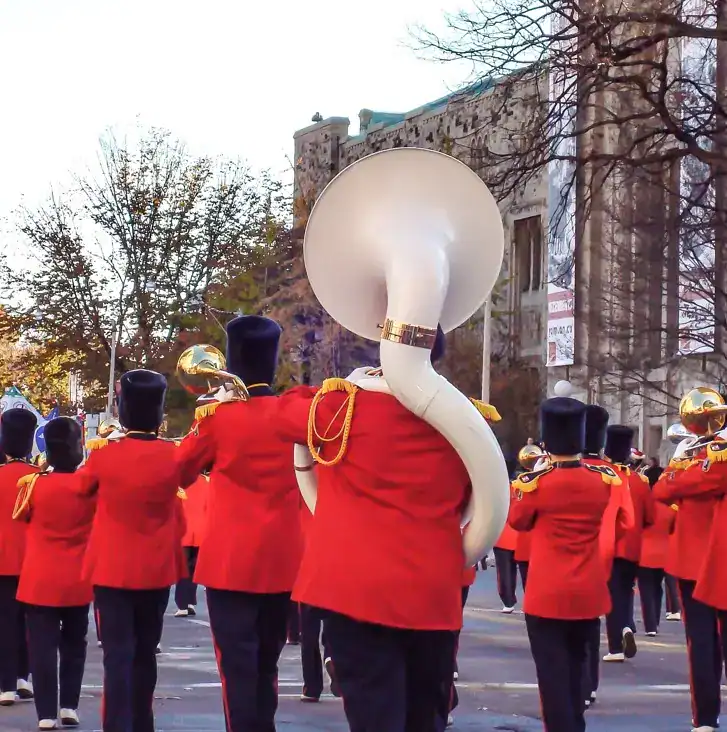 Banda de músicos en desfile con uniformes rojos y instrumentos, representando cultura y tradición, en una celebración en la calle con edificios y árboles de fondo.