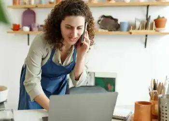 Emprendedora femenina en taller de cerámica, con computadora y teléfono móvil, resaltando el trabajo artesanal y digital en comunidad migrante en México. – SoyMigrante.com REVISTA