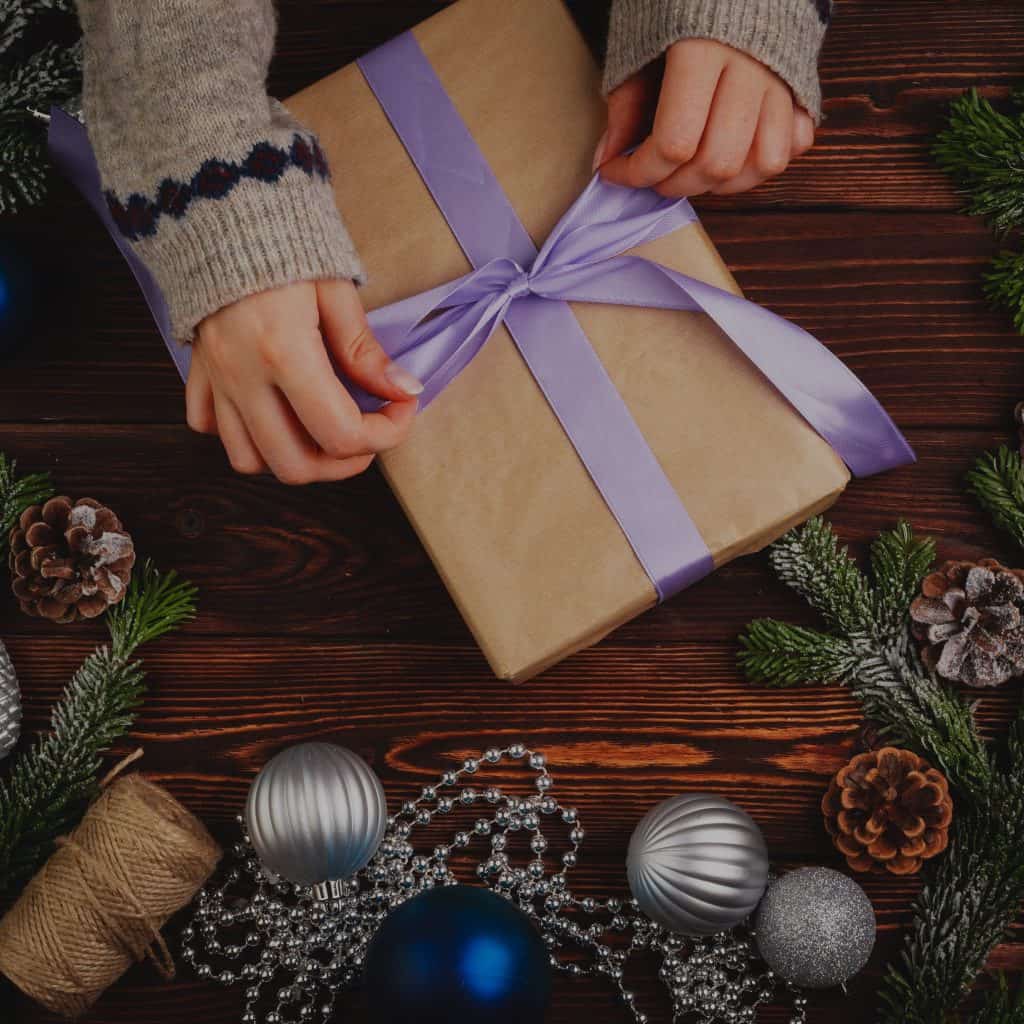 Regalo de Navidad envuelto con cintas moradas en mesa de madera decorada con bolas navideñas y piñas, celebración y tradición festiva. – SoyMigrante.com REVISTA