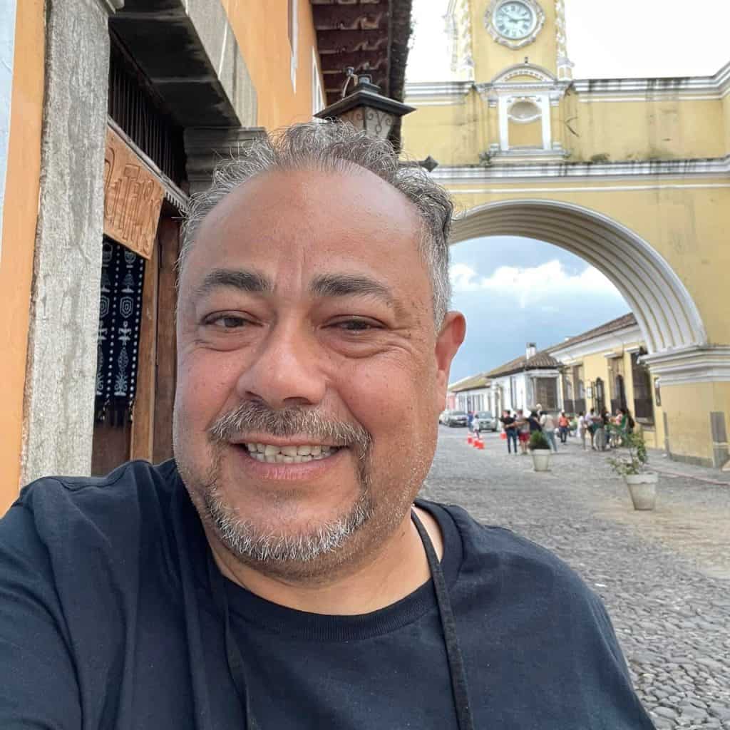 Retrato de un hombre sonriendo en una calle histórica, en un entorno cultural y turístico en Guatemala, destacando la importancia de la comunidad migrante en América Latina y enriqueciendo contenido para revista migrante. – SoyMigrante.com REVISTA
