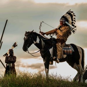 Guerrero indígena montado a caballo con plumas tradicionales en ceremonia cultural en la naturaleza. – SoyMigrante.com REVISTA