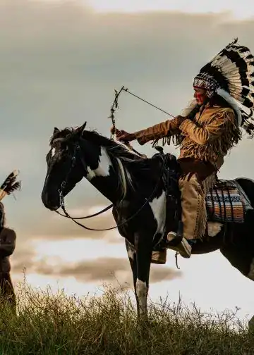 Guerrero indígena montado a caballo con plumas tradicionales en ceremonia cultural en la naturaleza. – SoyMigrante.com REVISTA