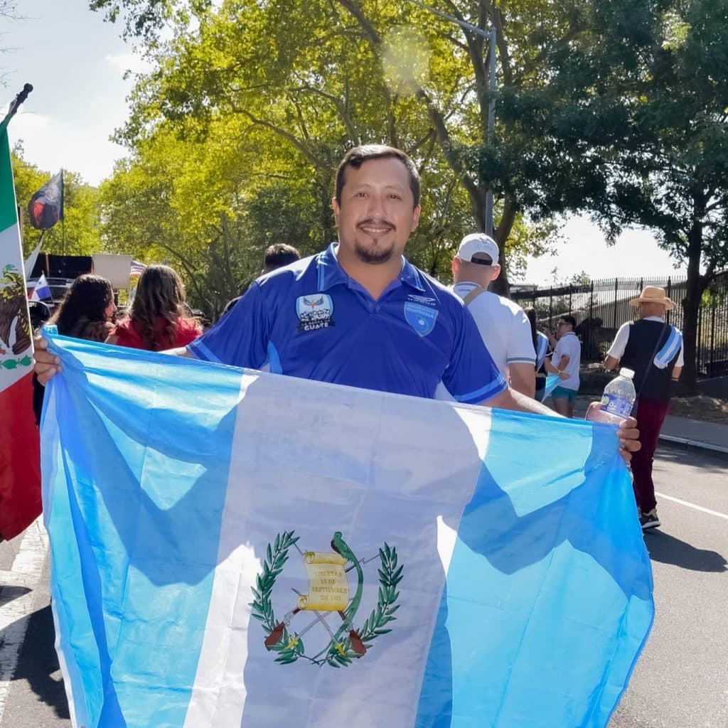 Guatemalteco con bandera en marcha patriótica, símbolo de orgullo y celebración nacional, participando en evento cultural y cívico. – SoyMigrante.com REVISTA