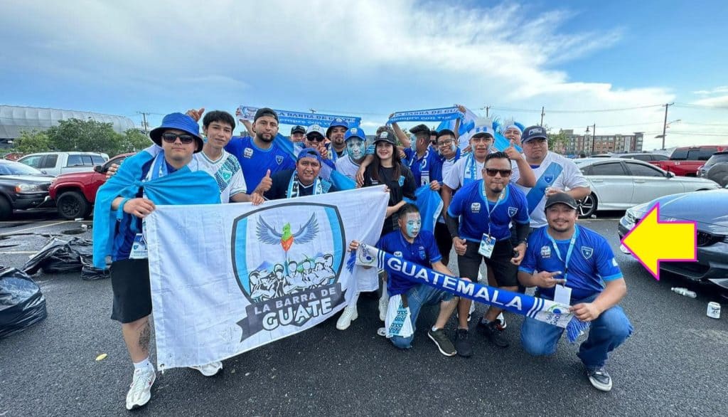 Varios aficionados de la selección de Guatemala en un evento deportivo, con banderas y camisetas del equipo, en un estacionamiento al aire libre celebrando el fútbol guatemalteco. – SoyMigrante.com REVISTA