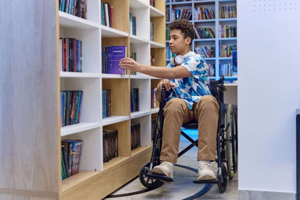 Joven en silla de ruedas explorando libros en una biblioteca, promoviendo inclusión, accesibilidad y diversidad en el aprendizaje y la cultura para migrantes y personas con discapacidad. – SoyMigrante.com REVISTA