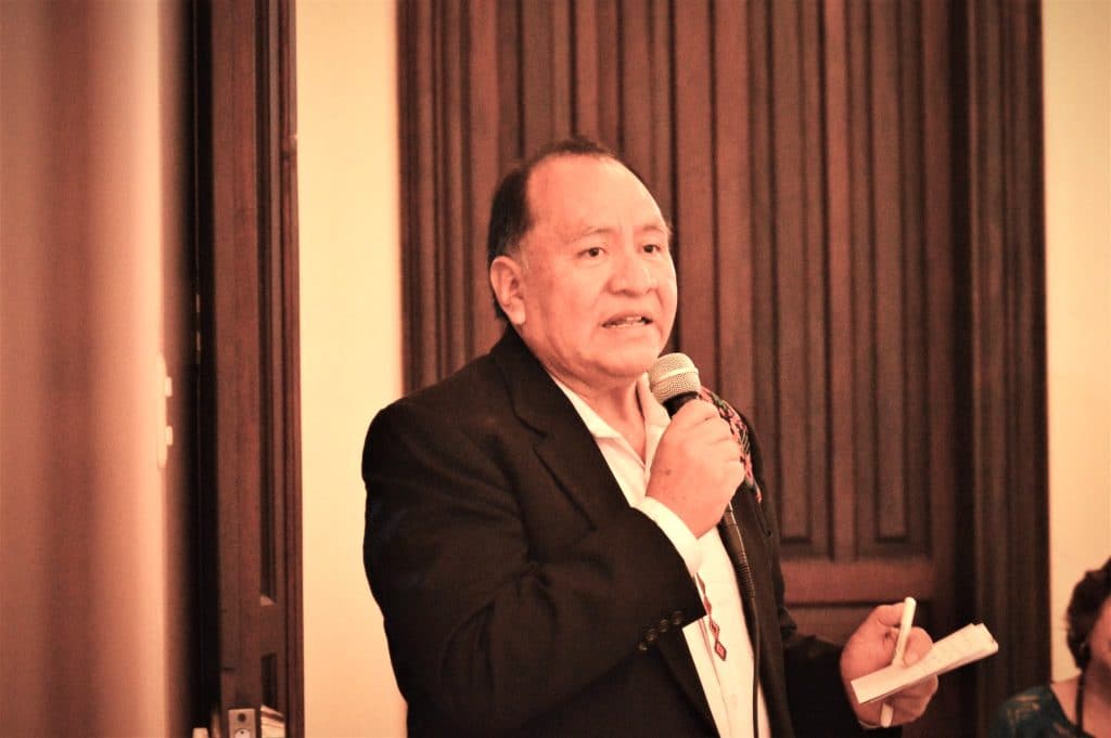 Hombre en conferencia, hablando con micrófono y portapapeles en mano, en evento de revista SoyMigrante.com REVISTA. – SoyMigrante.com REVISTA