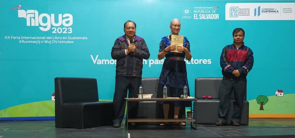 Biblioteca en evento cultural en Guatemala con personas en escenario, fondo azul con logos y texto informativo, varios asistentes participando en feria internacional del libro. – SoyMigrante.com REVISTA