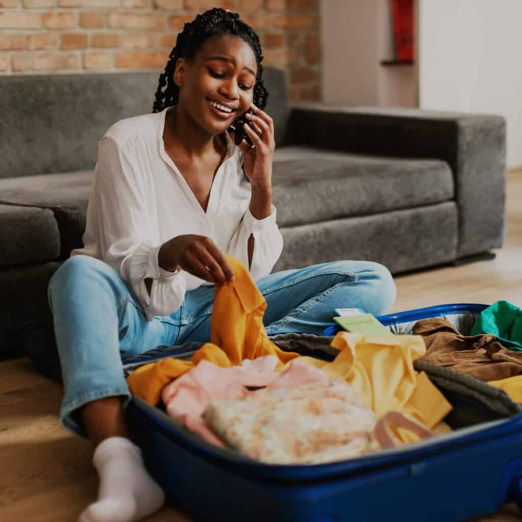 Viajera mujer hablando por teléfono mientras prepara maleta con ropa en casa. – SoyMigrante.com REVISTA