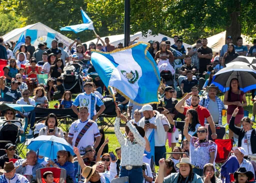 Banderas y símbolos de Guatemala en una celebración cultural llena de inmigrantes y comunidad en un parque, promoviendo la identidad y orgullo guatemalteco. – SoyMigrante.com REVISTA