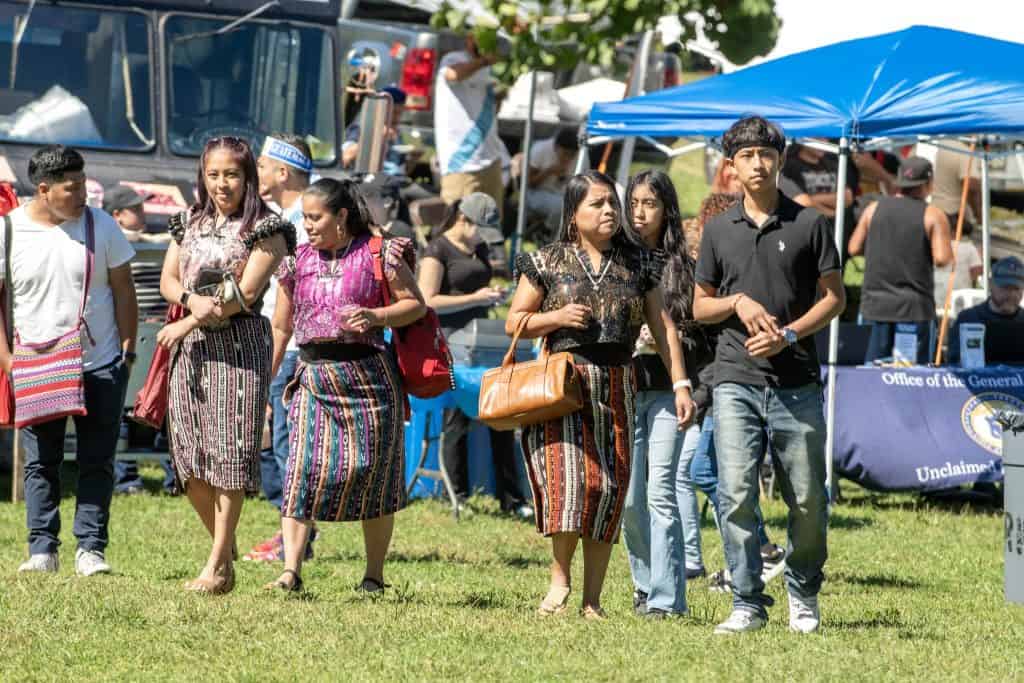 Personas migrantes vestidas con trajes tradicionales en evento cultural en un parque, participando en festividades de la comunidad migrante en Estados Unidos, resaltando identidad y orgullo cultural.