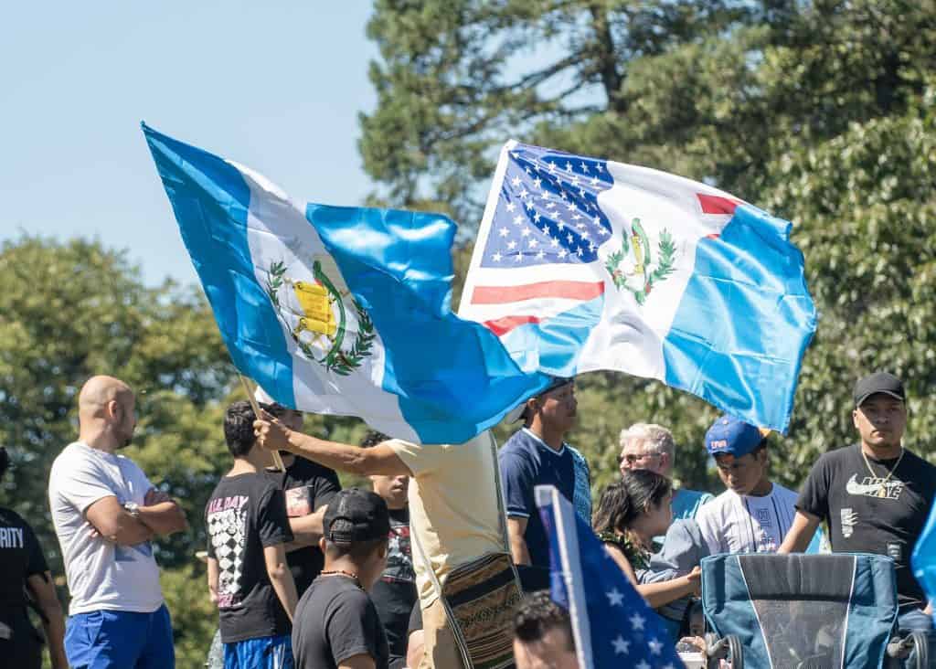 Banderas de Guatemala y Estados Unidos en un evento migrante, simbolizando la unión y comunidad de migrantes en un entorno al aire libre, con árboles y cielo azul de fondo. – SoyMigrante.com REVISTA