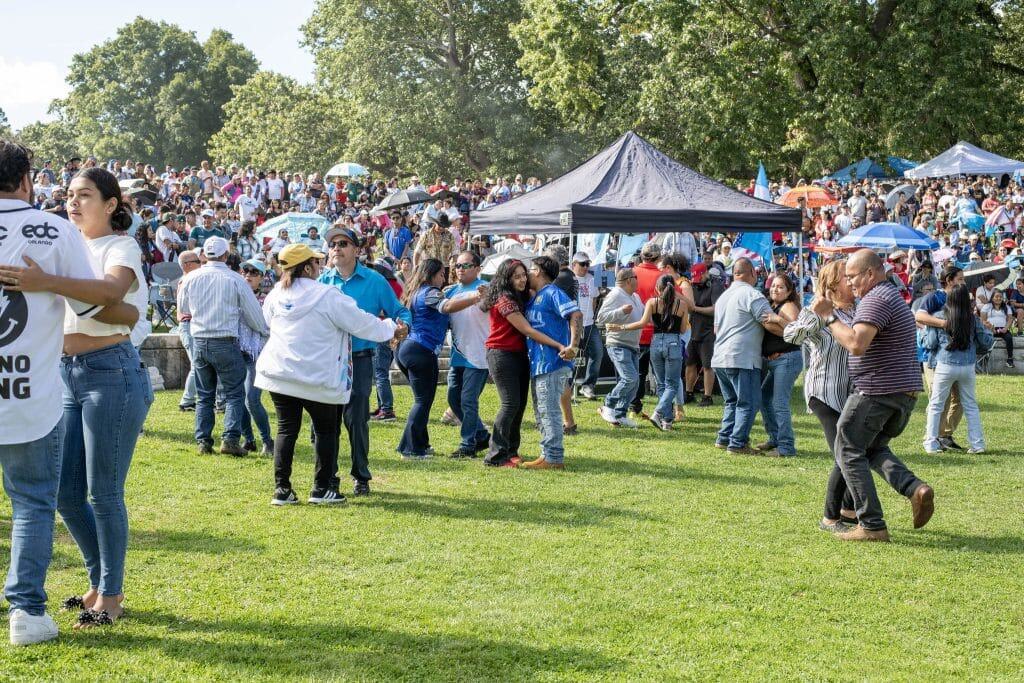 Celebración cultural y comunidad en evento de inmigrantes en un parque al aire libre en Estados Unidos. Personas bailan, conversan y disfrutan en un ambiente festivo.