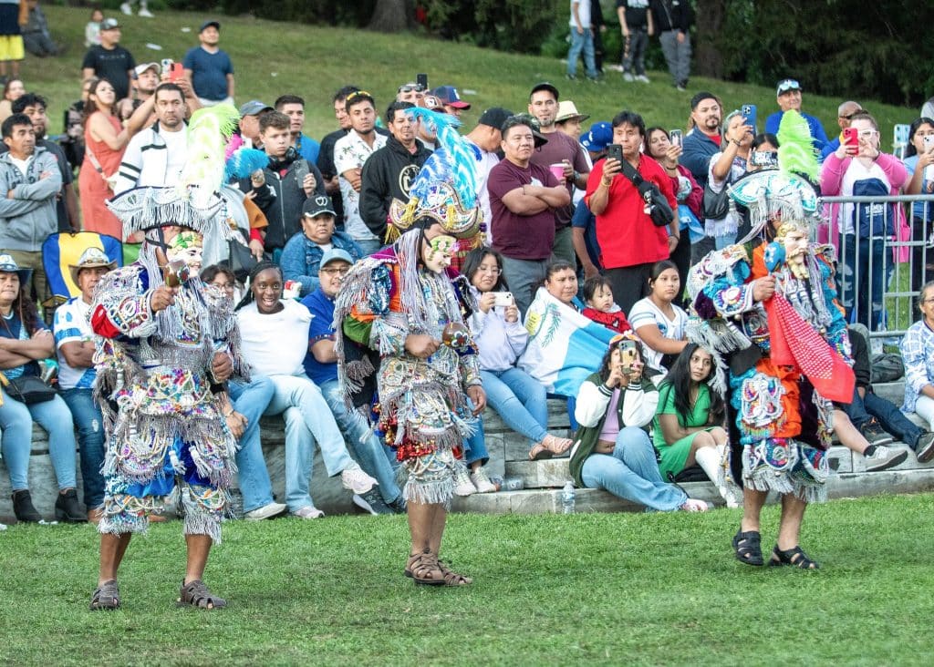 Coloridos bailarines tradicionales tlaxcaltecas en una celebración cultural en un parque, rodeados de un público observador y tomando fotos, resaltando la identidad y tradiciones mexicanas.