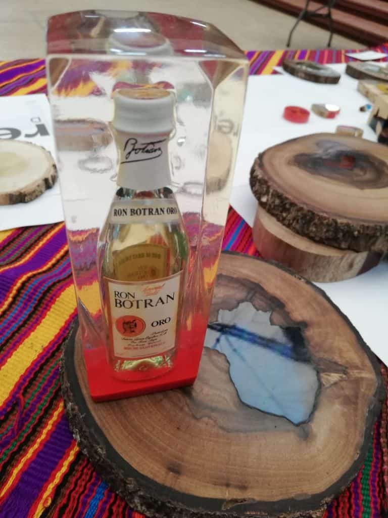 Ron Botran Oro botella de ron en exhibición en caja de acrílico con base roja, rodeada de madera decorativa y textiles de colores vibrantes, en un entorno que destaca la cultura y tradición del ron guatemalteco. – SoyMigrante.com REVISTA