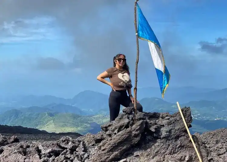 Campesina en la cima de un volcán con bandera de Guatemala, símbolo de lucha y orgullo indígena, en un paisaje volcánico y montañoso, en el contexto de la Revista SoyMigrante.com.