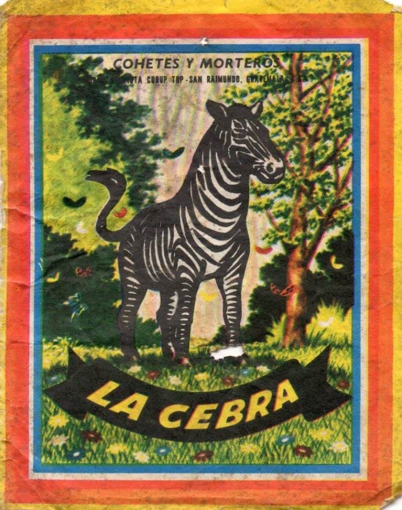 Zebra ilustración de una caja de cohetes y morteros en un entorno natural con árboles y flores, título "La Cebra". Enfoque en cultura, historia y tradiciones mexicanas. – SoyMigrante.com REVISTA