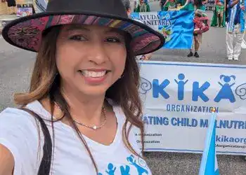 Niña en desfile cultural participando en evento de apoyo a la infancia y la nutrición en México, con grupo de personas en vestimenta tradicional y cartel de organización KIKA, promoviendo derechos infantiles.