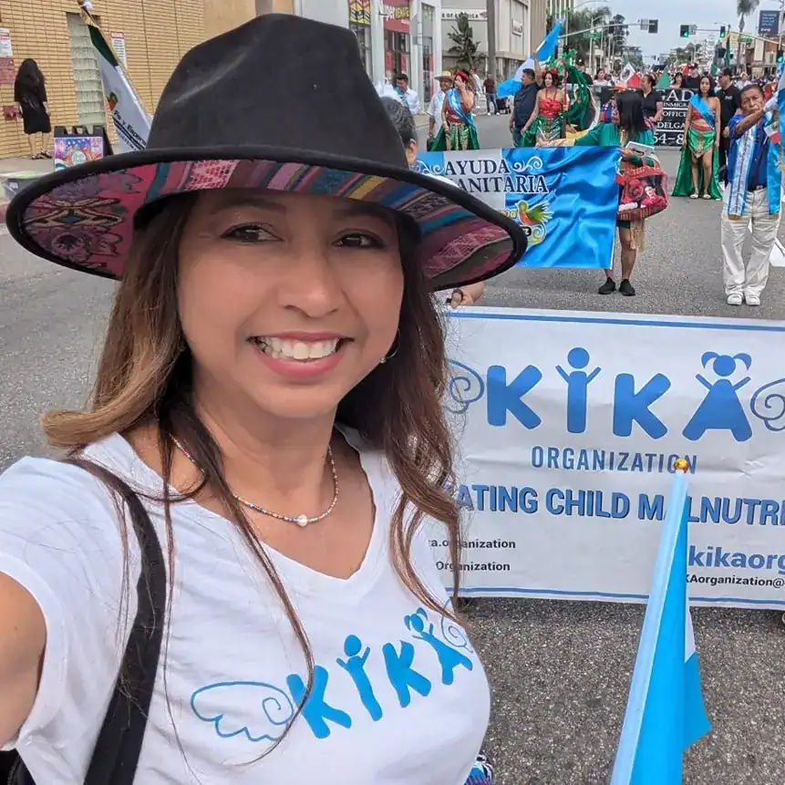 Niña en desfile cultural participando en evento de apoyo a la infancia y la nutrición en México, con grupo de personas en vestimenta tradicional y cartel de organización KIKA, promoviendo derechos infantiles.