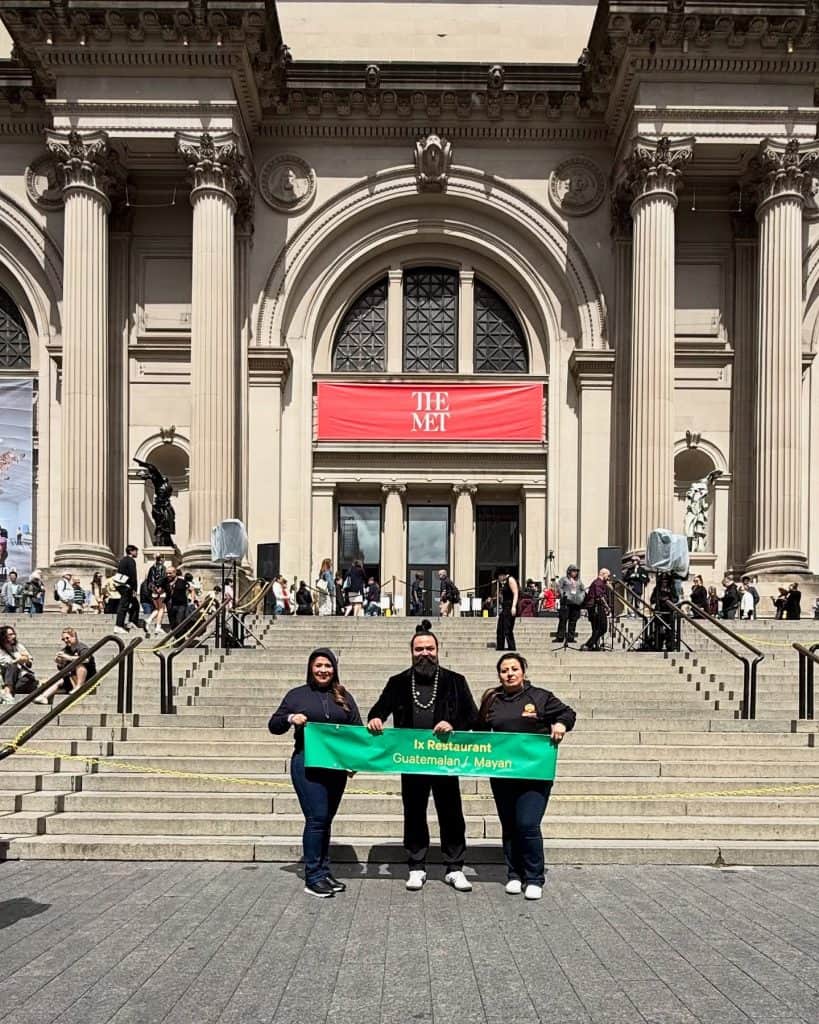 Restaurantes guatemaltecos en el exterior, promoviendo cultura y gastronomía en eventos internacionales en el MET de Nueva York. La imagen muestra a tres personas con una pancarta que dice "IX Restaurant Guatemalan / Mayan". – SoyMigrante.com REVISTA