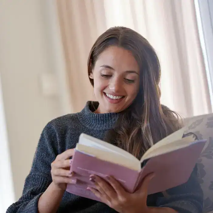 Joven mujer leyendo revista SoyMigrante.com REVISTA, con sonrisa y en interior, promoviendo información y cultura sobre migración y derechos. – SoyMigrante.com REVISTA