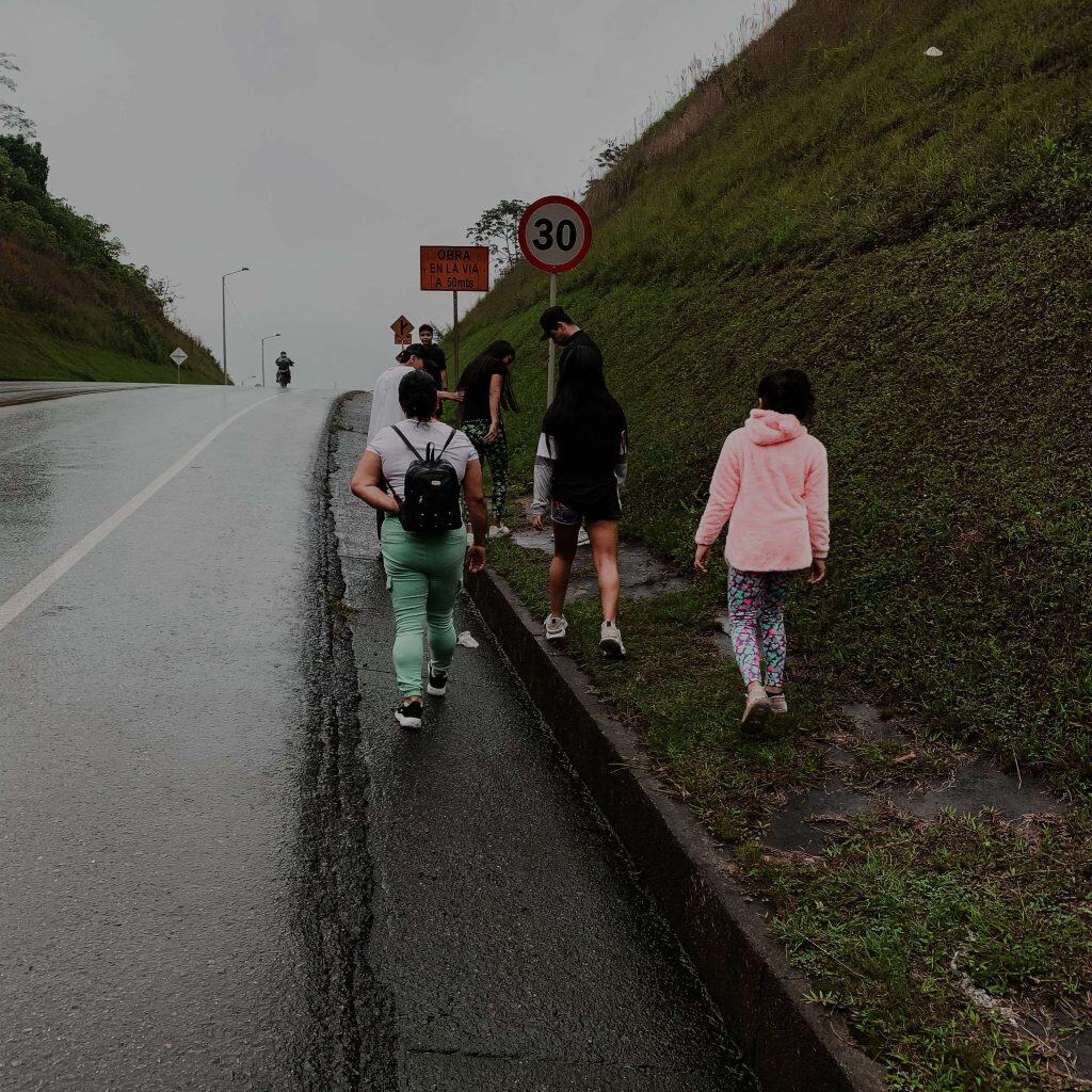 Camino con peatones en carretera lluviosa, señal de obra vial y límite de velocidad de 30 km/h, en un entorno natural y montañoso, en la publicación de SoyMigrante.com REVISTA. – SoyMigrante.com REVISTA
