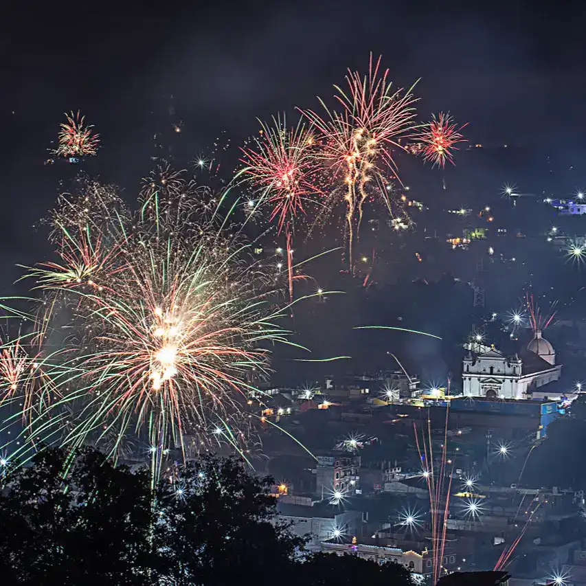 Fuegos artificiales en una celebración nocturna en una ciudad con iglesia histórica de fondo, protagonizando la portada de SoyMigrante.com REVISTA. – SoyMigrante.com REVISTA