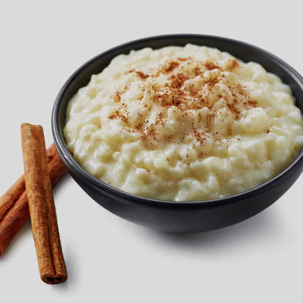 Suave image de un delicioso arroz con leche en un bol negro, espolvoreado con canela y acompañado de canelas en rama, representando la cultura gastronómica latinoamericana y la tradición culinaria. – SoyMigrante.com REVISTA