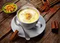 Tazón de crema de elote y canela en plato blanco, acompañada de maíz y canela en un fondo de madera rustic; gastronomía tradicional mexicana, bebida caliente, sabor auténtico.