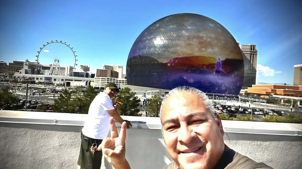 Rostro de un hombre sonriendo y haciendo señal de paz, con una vista de Las Vegas y la Bola LED en el fondo, en la azotea de un edificio, promoviendo la comunidad migrante y el turismo en Estados Unidos. – SoyMigrante.com REVISTA