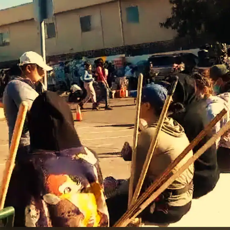 Personas migrantes en fila en el exterior, participando en actividad comunitaria o social en un ambiente urbano al aire libre. – SoyMigrante.com REVISTA