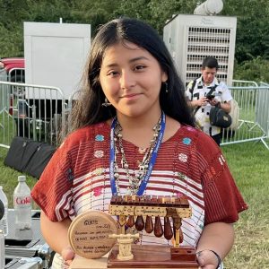 Joven indígena con premio en feria cultural, usando vestimenta tradicional, con la inscripción "Guate", en evento de reconocimiento a jóvenes talentos en Guatemala, promoviendo la cultura y la identidad indígena. – SoyMigrante.com REVISTA