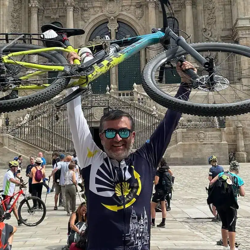 Bicicleta de montaña levantada en el aire frente a una iglesia histórica, ciclista sonriente con gafas deportivas y bandera, grupo de ciclistas en la plaza, deporte y aventura en espacios urbanos y culturales. – SoyMigrante.com REVISTA