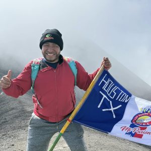 Explorador de montaña en la cima, mostrando entusiasmo con bandera de Houston TX, foto en la cumbre, aventura, inmigrante, comunidad, superación, pasión por la vida. – SoyMigrante.com REVISTA