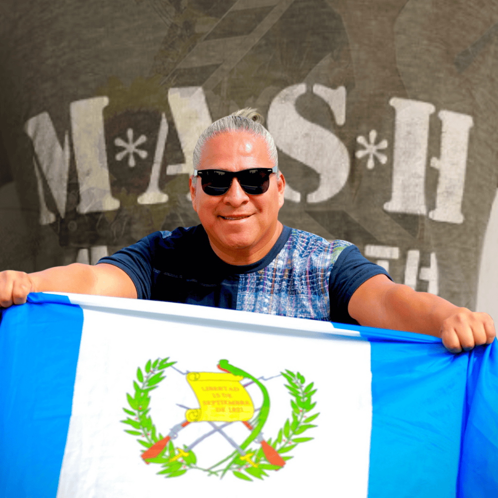 Joven con gafas oscuras sosteniendo la bandera de Guatemala frente a un cartel de SoyMigrante.com REVISTA. – SoyMigrante.com REVISTA