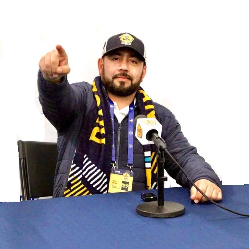 Solidario líder de la comunidad migrante en una entrevista, usando bufanda y gorra con logotipo, en un entorno de prensa y comunicación escrita centrada en temas de migrantes y derechos sociales.