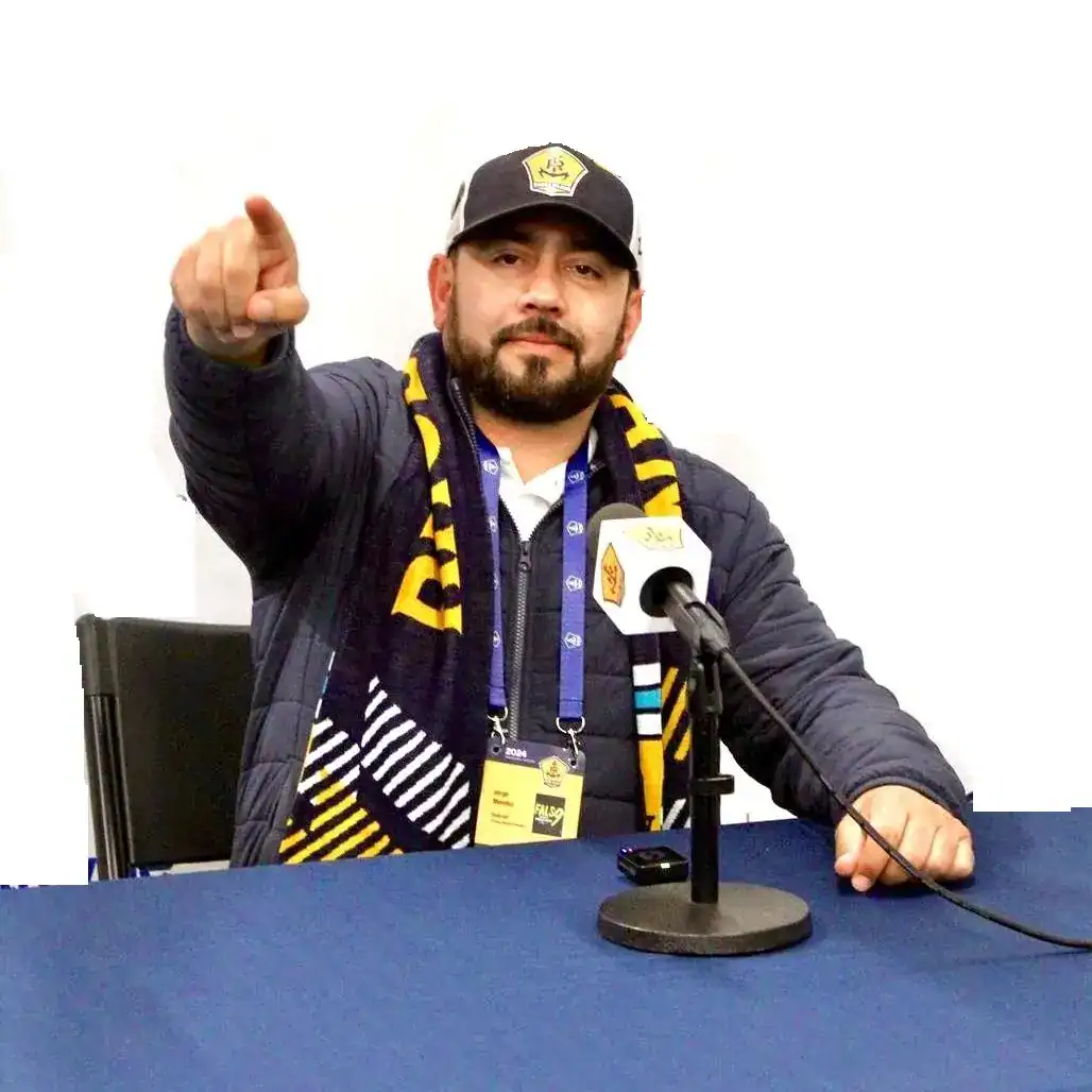 nacho mendez migrante guatemalteco 2 – SoyMigrante.com REVISTA Solidario líder de la comunidad migrante en una entrevista, usando bufanda y gorra con logotipo, en un entorno de prensa y comunicación escrita centrada en temas de migrantes y derechos sociales.