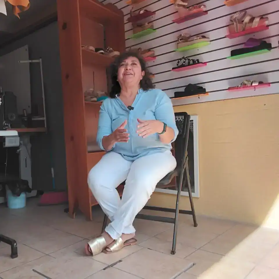 Zapatos de tacón dorados y pantalones blancos casuales con blusa azul claro, sentada en una silla en una tienda de zapatos con exhibición de sandalias y gafas en el fondo.