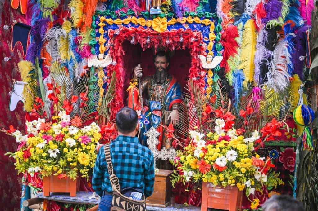 Colorido altar de Día de Muertos con flores y advocaciones religiosas, símbolo de la cultura mexicana y tradición ancestral, decorado con vibrantas plumas y figuras religiosas. – SoyMigrante.com REVISTA