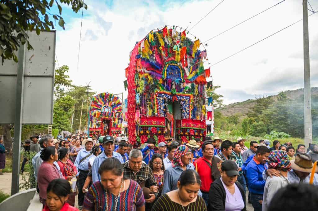 Cola de personas en celebración tradicional con grandes carros alegóricos adornados con colores vibrantes en un entorno rural en México. – SoyMigrante.com REVISTA
