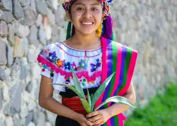 Colombia mujer indígena en vestimenta tradicional con flores y frutas coloridas, en una pose alegre y cultural. – SoyMigrante.com REVISTA